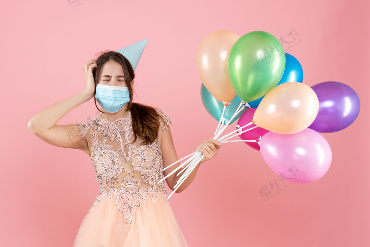生日戴着派对帽和医用面罩的派对女孩闭上眼睛 手里拿着粉色的彩色气球举行派对女孩派对