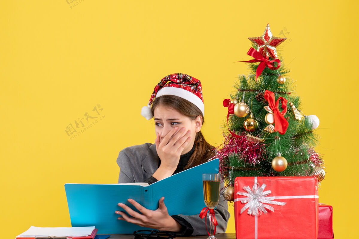 女孩前视图吓坏了戴圣诞帽的女孩坐在桌旁拿着文件圣诞树和礼物鸡尾酒桌子工作害怕的女孩