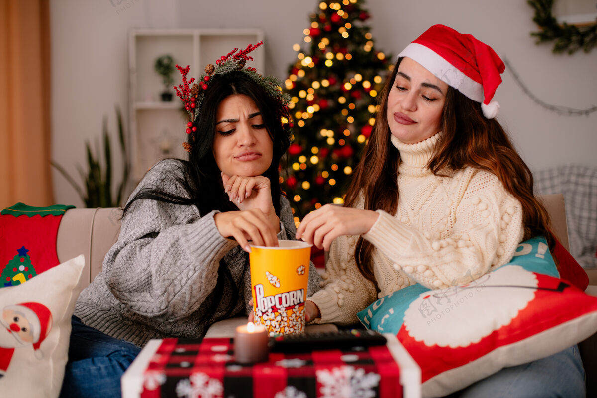 坐带着圣诞帽和冬青花环的漂亮女孩在家里吃东西 看着放在扶手椅上的爆米花桶年轻时间家