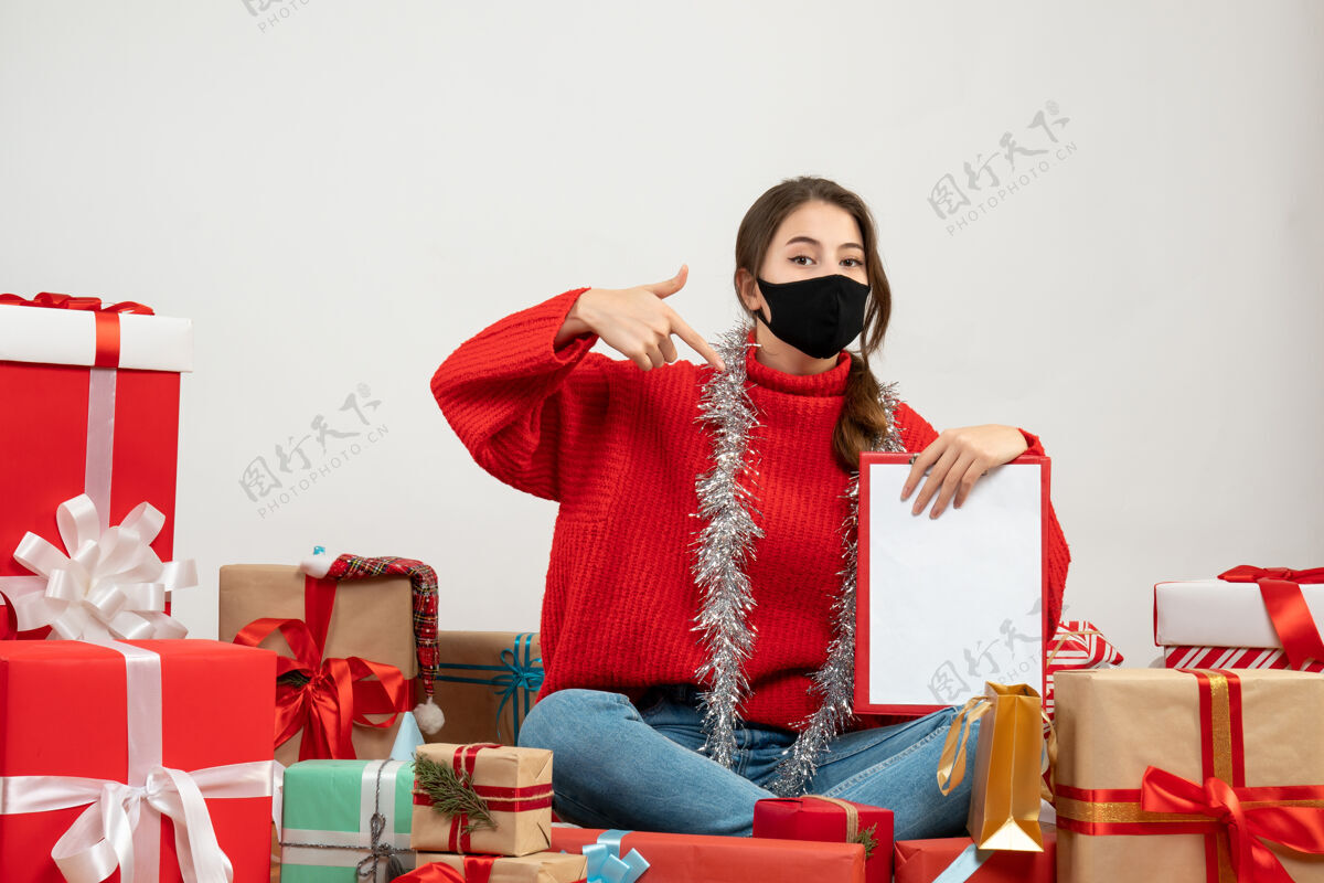 年轻女孩穿着红色毛衣的年轻女孩展示文件坐在礼物旁边 白色的面具上戴着黑色的面具女孩礼物圣诞节