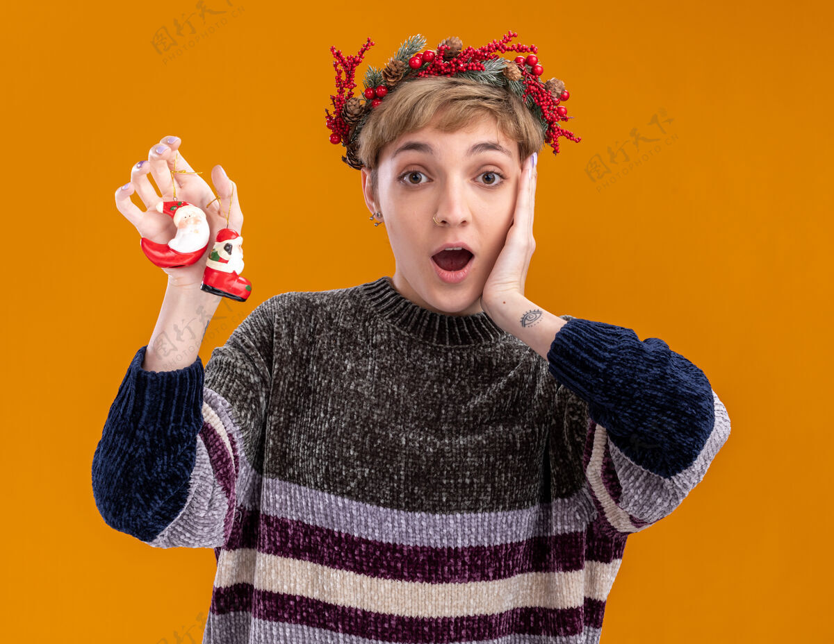 穿着令人印象深刻的年轻漂亮女孩戴着圣诞花环手持圣诞老人的圣诞饰品手放在脸上隔离在橙色的墙上圣诞老人漂亮花环