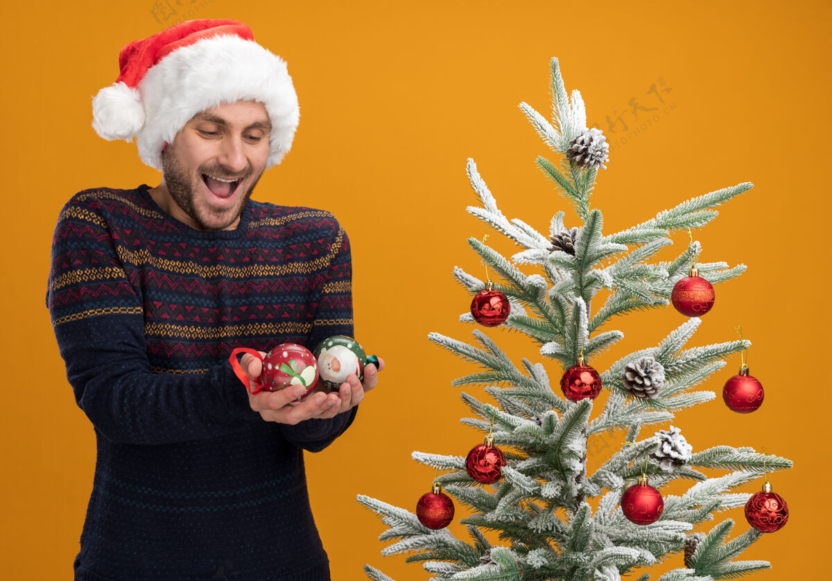 男人令人印象深刻的年轻白种人戴着圣诞帽站在装饰圣诞树附近拿着圣诞饰品看橙色墙上孤立印象深刻戴着圣诞节