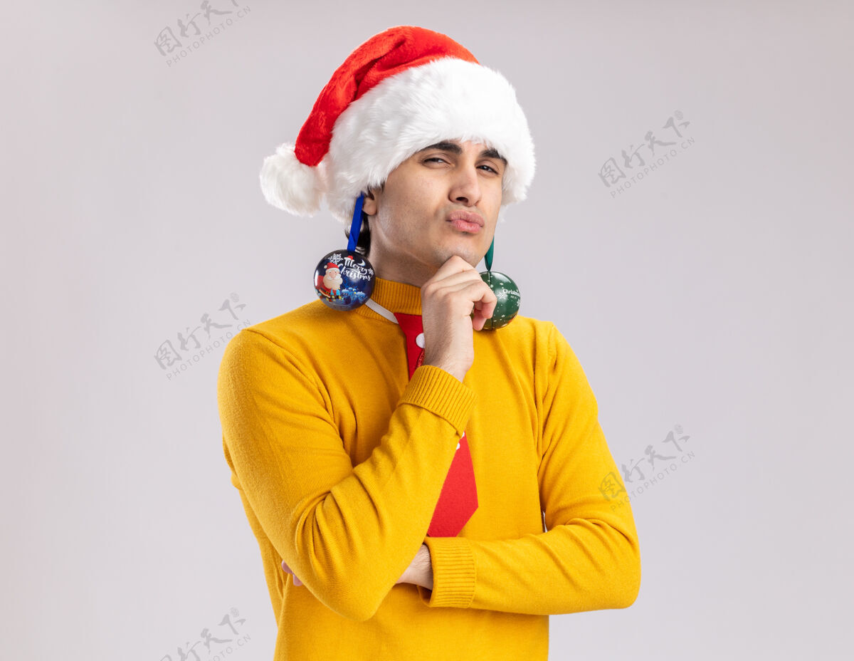 高领毛衣穿着黄色高领毛衣的年轻人和耳朵上戴着圣诞球的圣诞老人站在白色背景上 带着怀疑的表情看着摄像机年轻人舞会耳朵