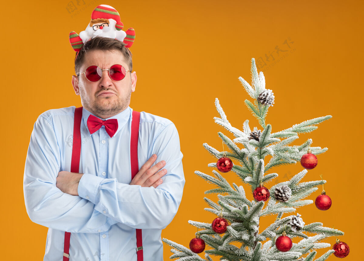 圣诞老人不高兴的年轻人穿着吊带领结在边缘与圣诞老人和红色眼镜站在旁边的圣诞树看着相机与手臂交叉在胸前橙色背景吊带不高兴领带
