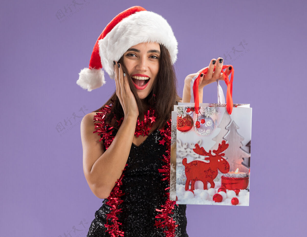 年轻快乐的年轻漂亮的女孩戴着圣诞帽 脖子上戴着花环 在镜头前拿出礼品袋 手放在脸颊上 在紫色的背景下与世隔绝女孩放脖子
