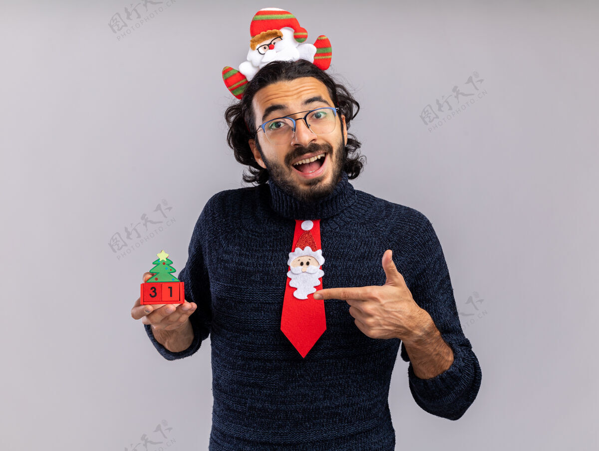 穿快乐的年轻帅哥戴着圣诞领带 戴着发箍 指着隔离在白墙上的圣诞玩具圣诞节快乐帅气