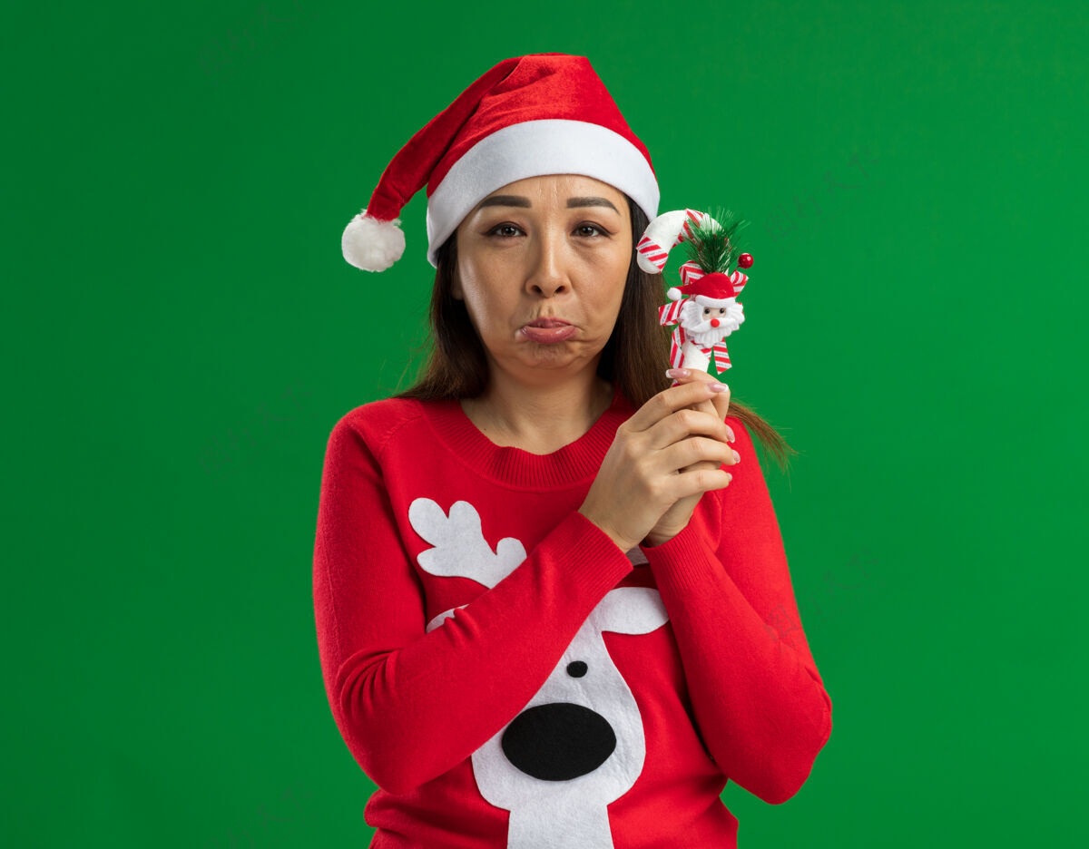 Pursing心烦意乱的年轻女子戴着圣诞老人帽 穿着红色毛衣 手持圣诞糖果手杖 看着相机 愁眉苦脸地撅着嘴唇站在绿色的背景上圣诞老人手杖嘴唇
