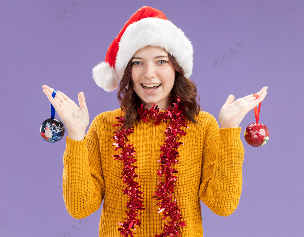 脖子快乐的年轻斯拉夫女孩 戴着圣诞帽 脖子上戴着花环 手拿玻璃球饰品 紫色背景上有复制空间球快乐圣诞老人