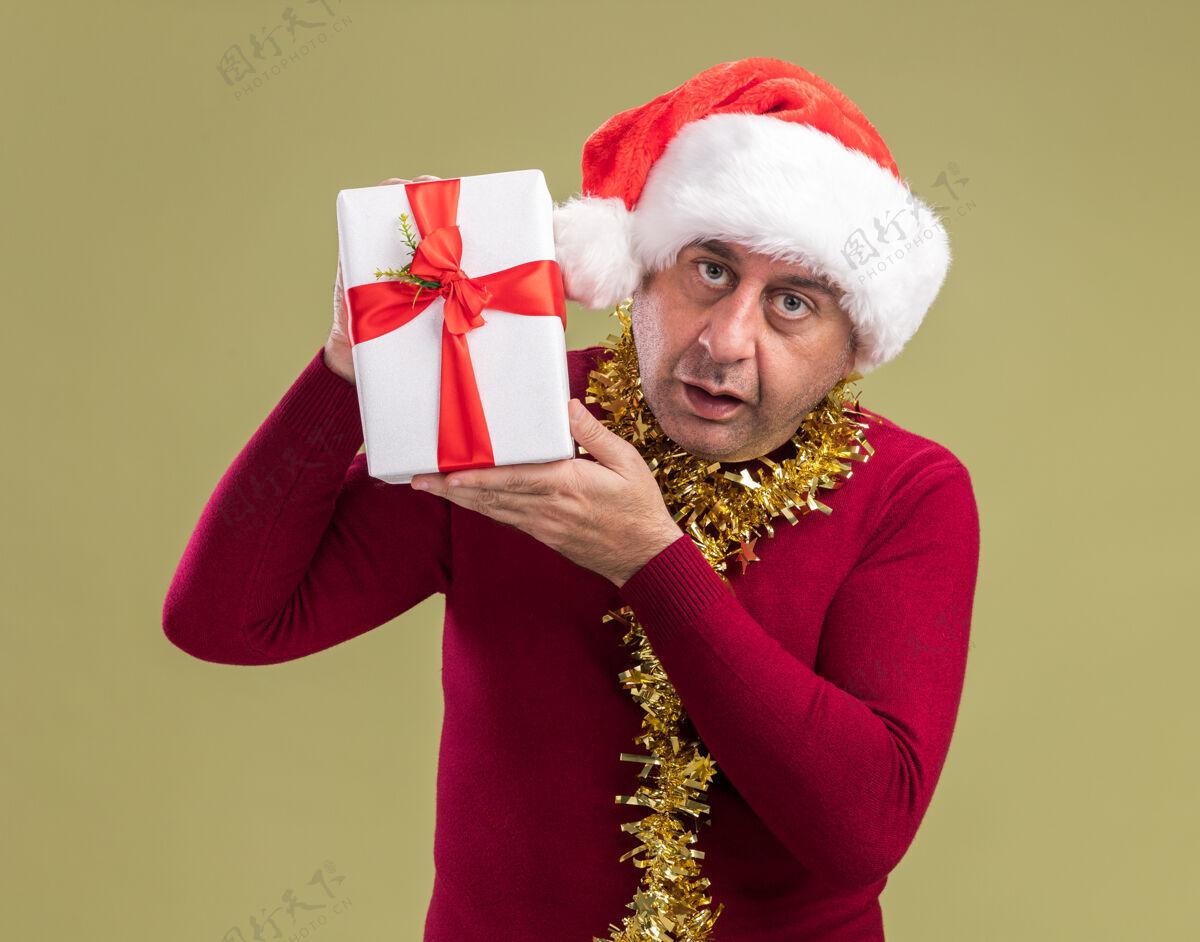 站着中年男子戴着圣诞老人帽子 脖子上戴着金属丝 手里拿着圣诞礼物 站在绿色的背景上困惑地看着相机礼物困惑金属片
