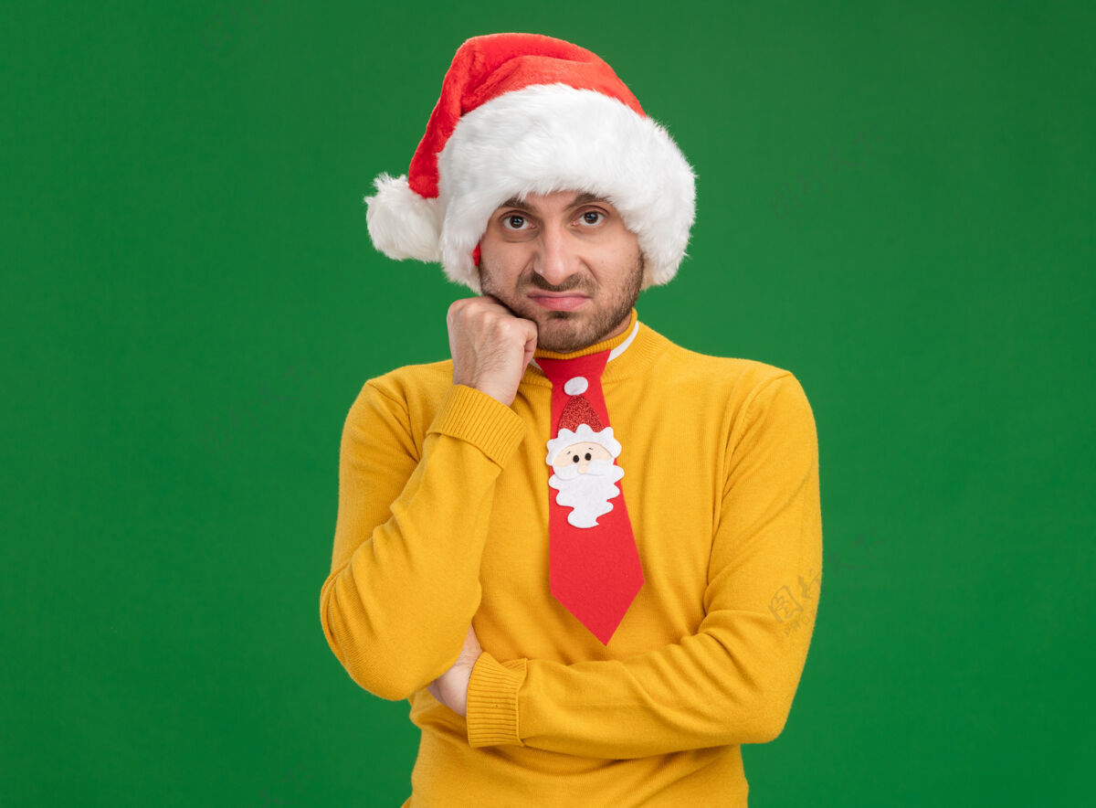 相机戴着圣诞帽 打着领带 手放在脸上 看着隔离在绿色背景下 有复印空间的相机 一个未经许可的白人年轻人年轻手领带