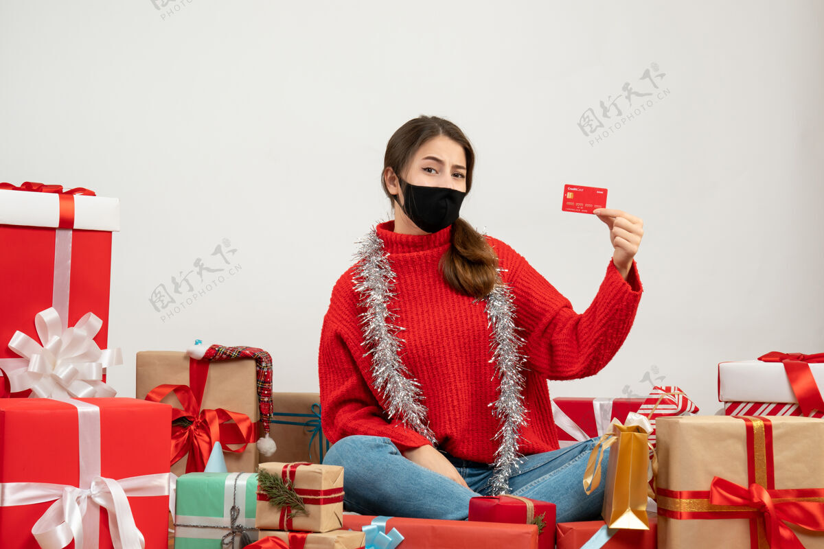 坐着穿着红色毛衣 戴着黑色面具 拿着信用卡的年轻女孩围坐在白色的礼物上服装年轻女孩圣诞节