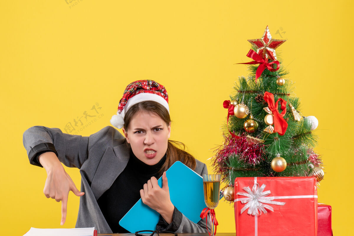 树前视图戴圣诞帽的年轻女孩坐在桌旁愤怒地表演圣诞树和礼物鸡尾酒鸡尾酒女孩表演