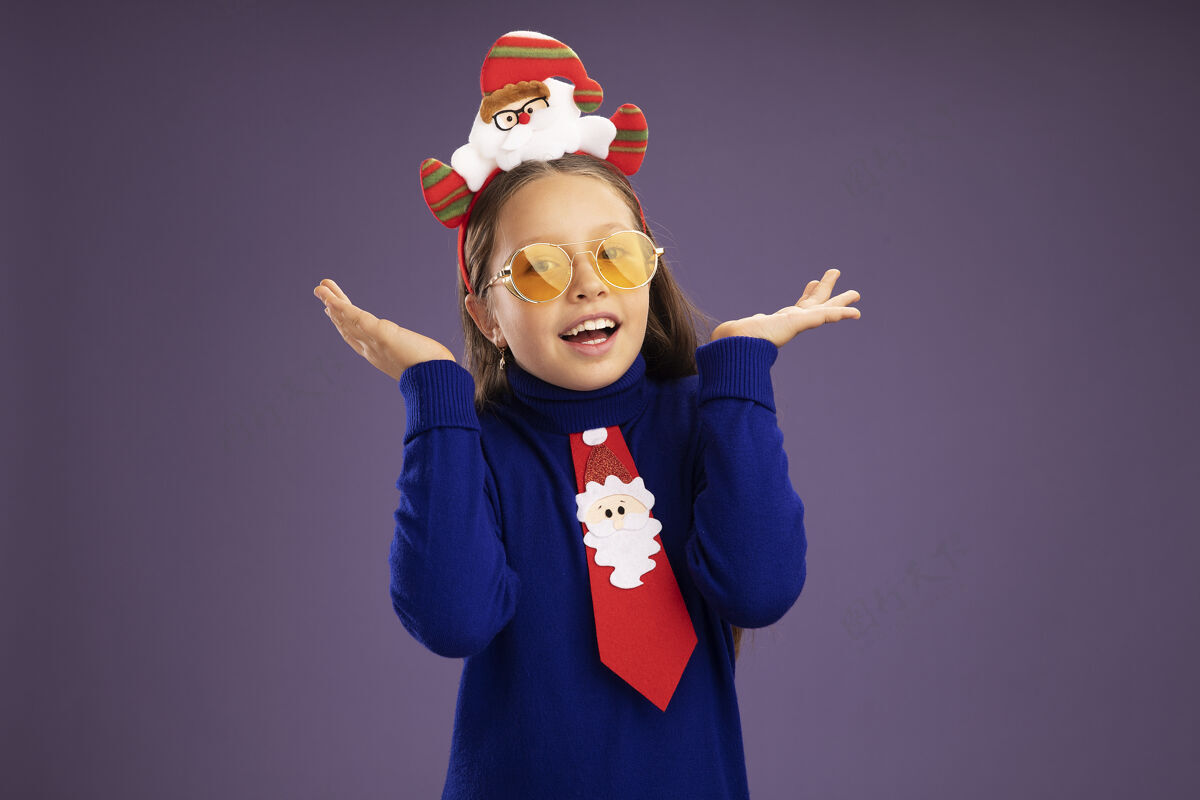 领带小女孩穿着蓝色高领毛衣 系着红色领带 头上戴着有趣的圣诞戒指 站在紫色的背景上 开心而兴奋地看着相机头兴奋手臂