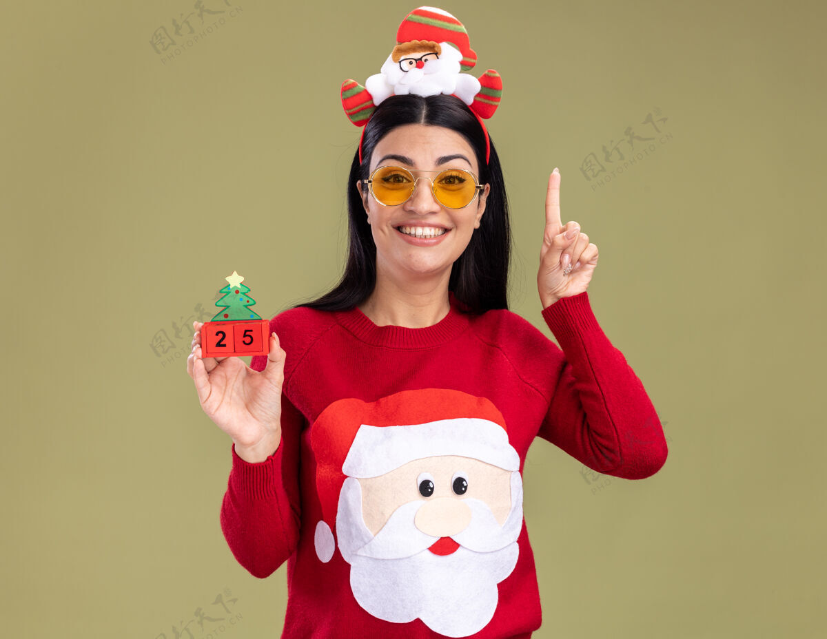 年轻微笑着的年轻白人女孩戴着圣诞老人的头带和带眼镜的毛衣拿着圣诞树玩具和日期看着相机指着橄榄绿背景上孤立眼镜圣诞树女孩
