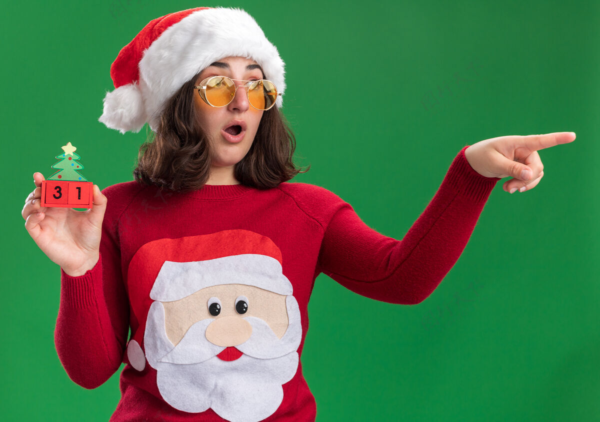 绿色穿着圣诞毛衣的小女孩戴着圣诞帽戴着眼镜拿着玩具方块和新年约会看立方体戴着
