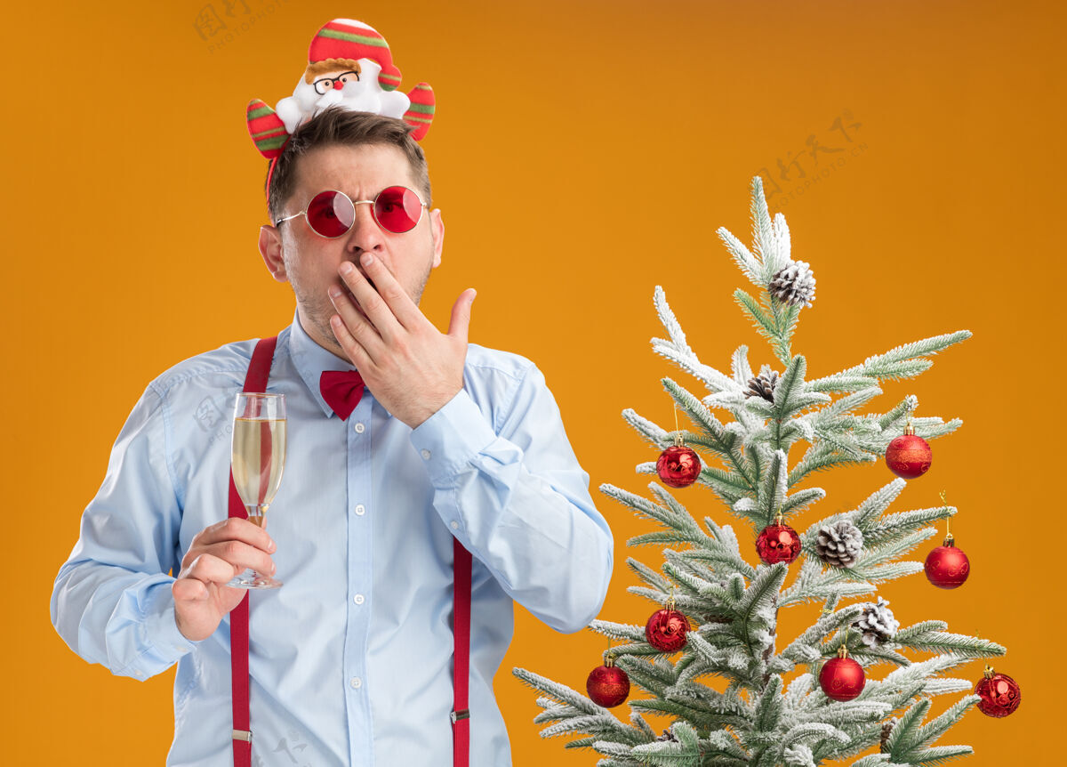 轮辋戴着吊带领结的年轻人站在圣诞树旁 手里拿着一杯香槟吊带快乐抱着