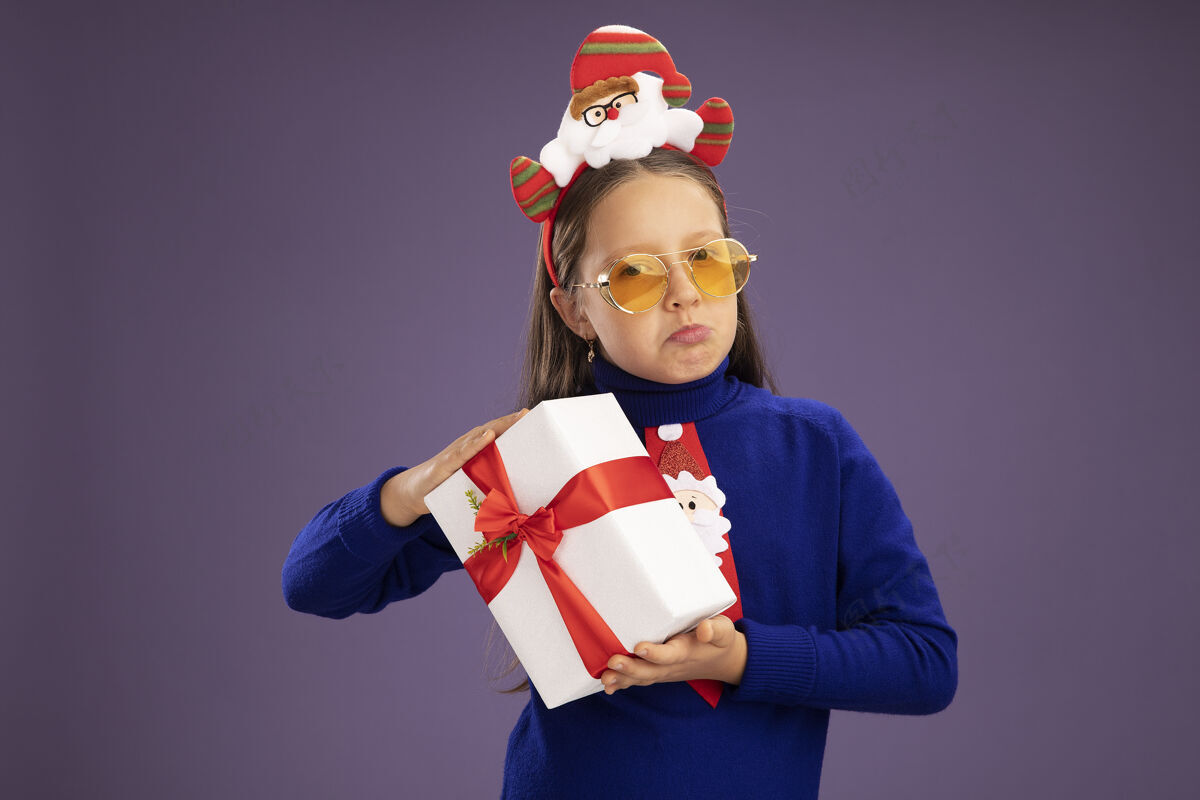 圣诞节小女孩穿着蓝色高领毛衣 打着红色领带 头上戴着有趣的圣诞戒指 手里拿着礼物 看着相机 脸上带着悲伤的表情 噘着嘴唇站在紫色的背景上领带抱着小