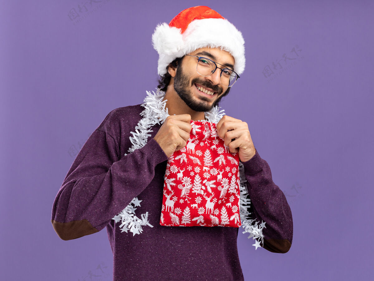年轻微笑的年轻帅哥戴着圣诞帽 脖子上戴着花环 手里拿着蓝色背景的圣诞包包圣诞花环