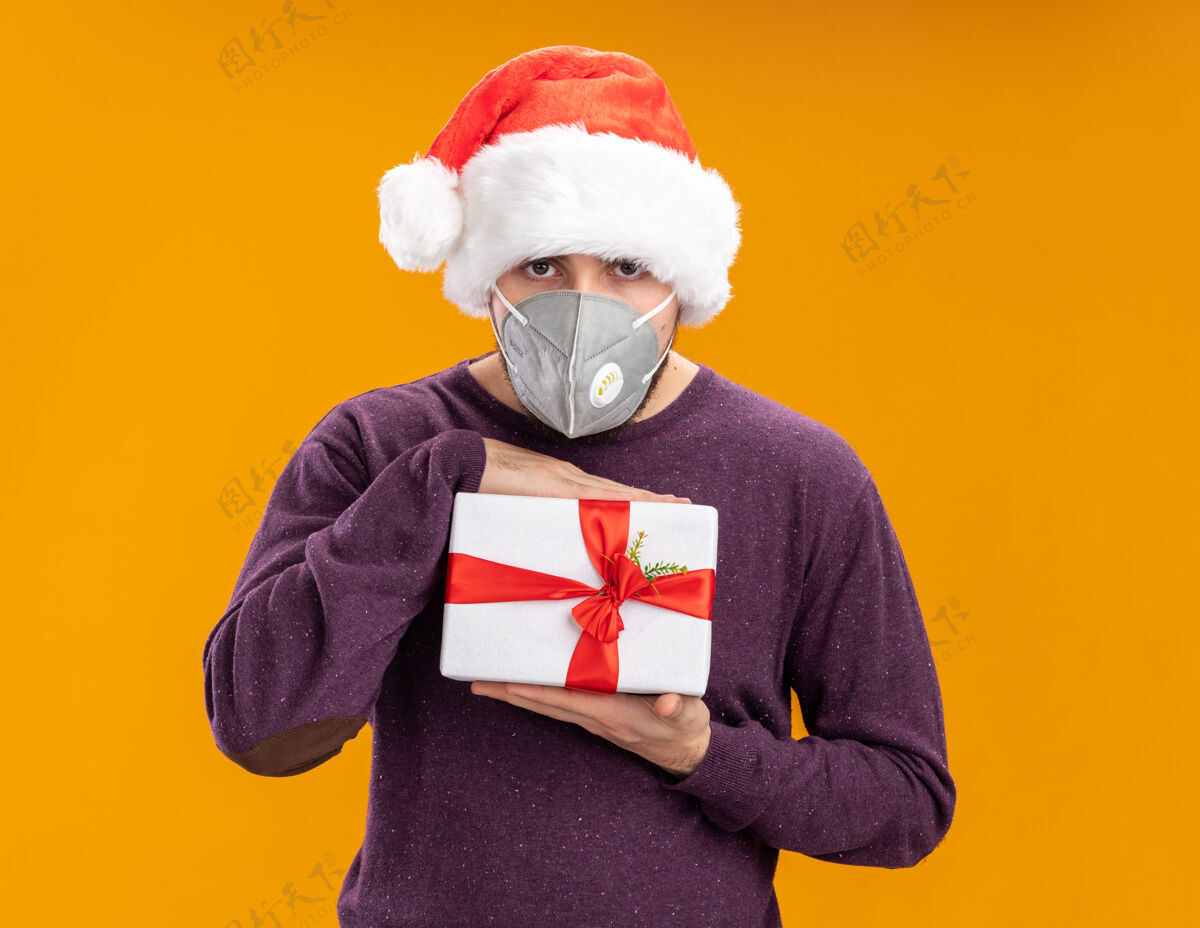 礼物穿着紫色毛衣 戴着圣诞帽 戴着护面面具的年轻人拿着礼物 严肃的脸站在橙色背景下看着摄像机脸圣诞老人年轻人