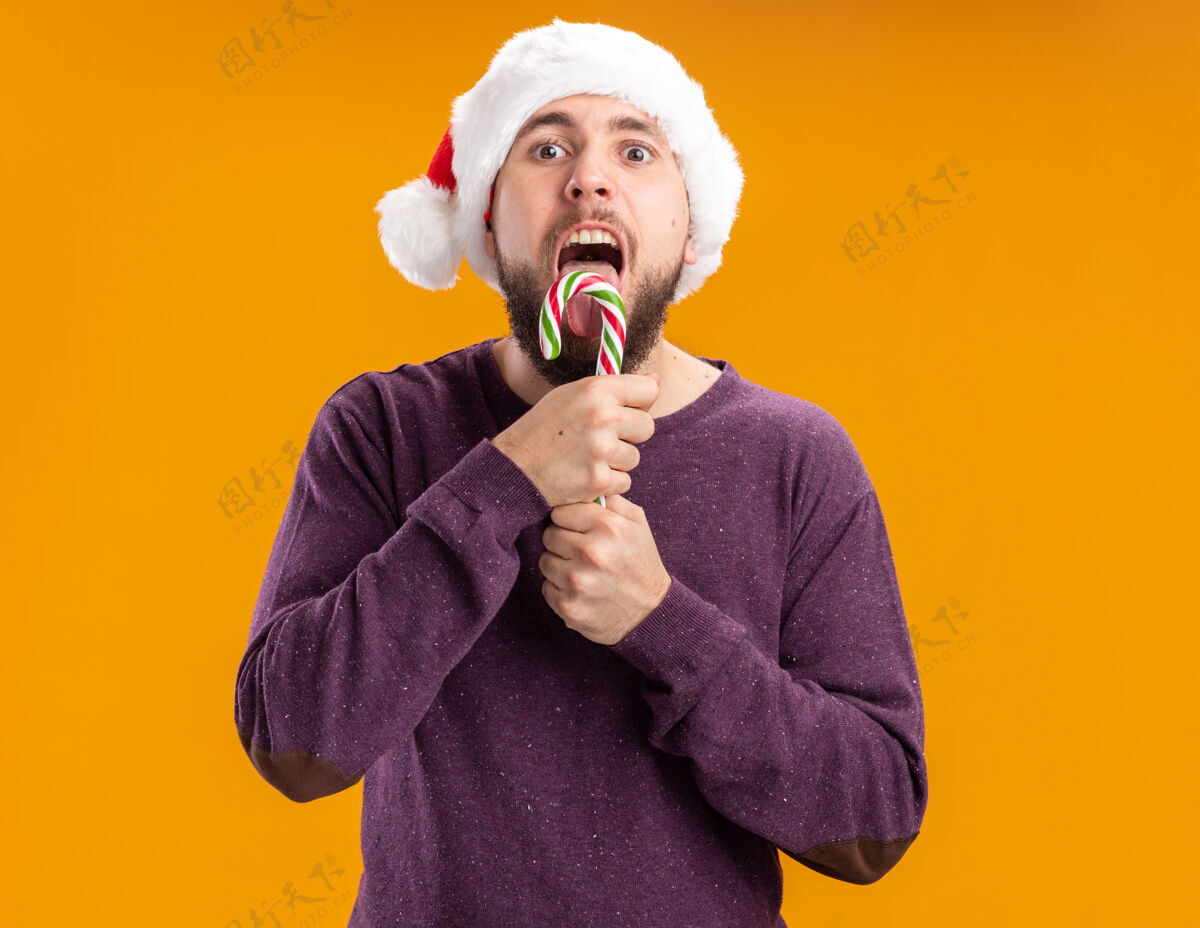 年轻身穿紫色毛衣 戴圣诞帽 手持糖果棒的年轻人站在橙色的背景下 要去品尝它的快乐和快乐圣诞老人毛衣糖果