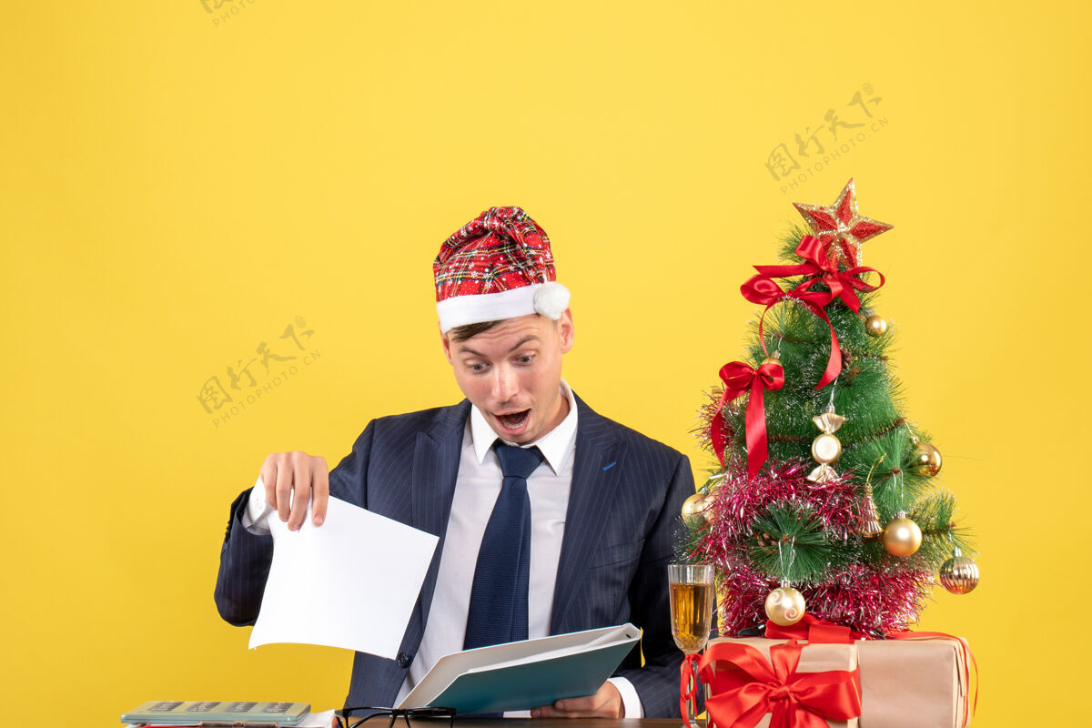 圣诞节商务人员坐在圣诞树旁的桌子旁检查文件和黄色墙上的礼物的前视图检查行政人员坐着