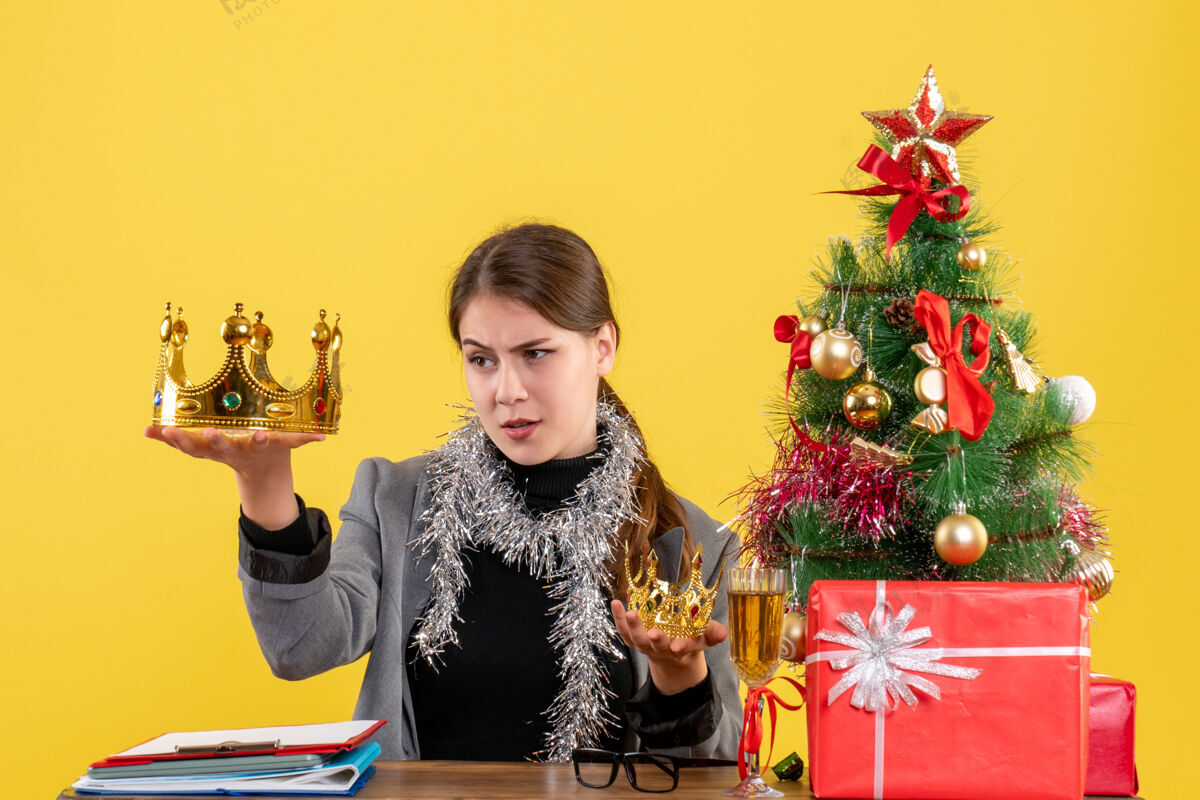 节日正面图年轻女孩手持大大小小的皇冠 查看大大的圣诞树和礼物鸡尾酒节日桌子礼物