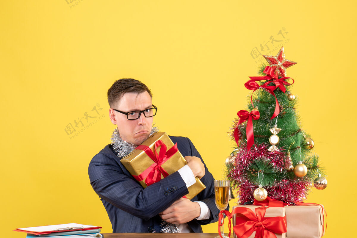 快乐前视图焦躁不安的男子紧紧地拿着他的礼物坐在圣诞树旁边的桌子上 黄色墙上的礼物复制空间礼物微笑礼物