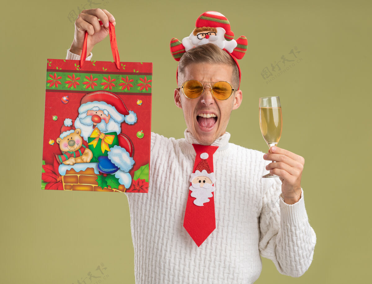 相机戴着圣诞老人头带和领带的年轻帅哥看着相机 手里拿着一杯香槟 举起圣诞礼包 闭着眼睛尖叫着 背景是橄榄绿圣诞节年轻提高