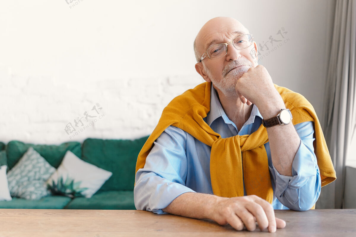 老人戴着长方形眼镜 肩上系着毛衣的资深秃头心理学家坐在家里办公室的空木桌旁 等待着他的委托人 有着若有所思的表情祖父眼镜桌子