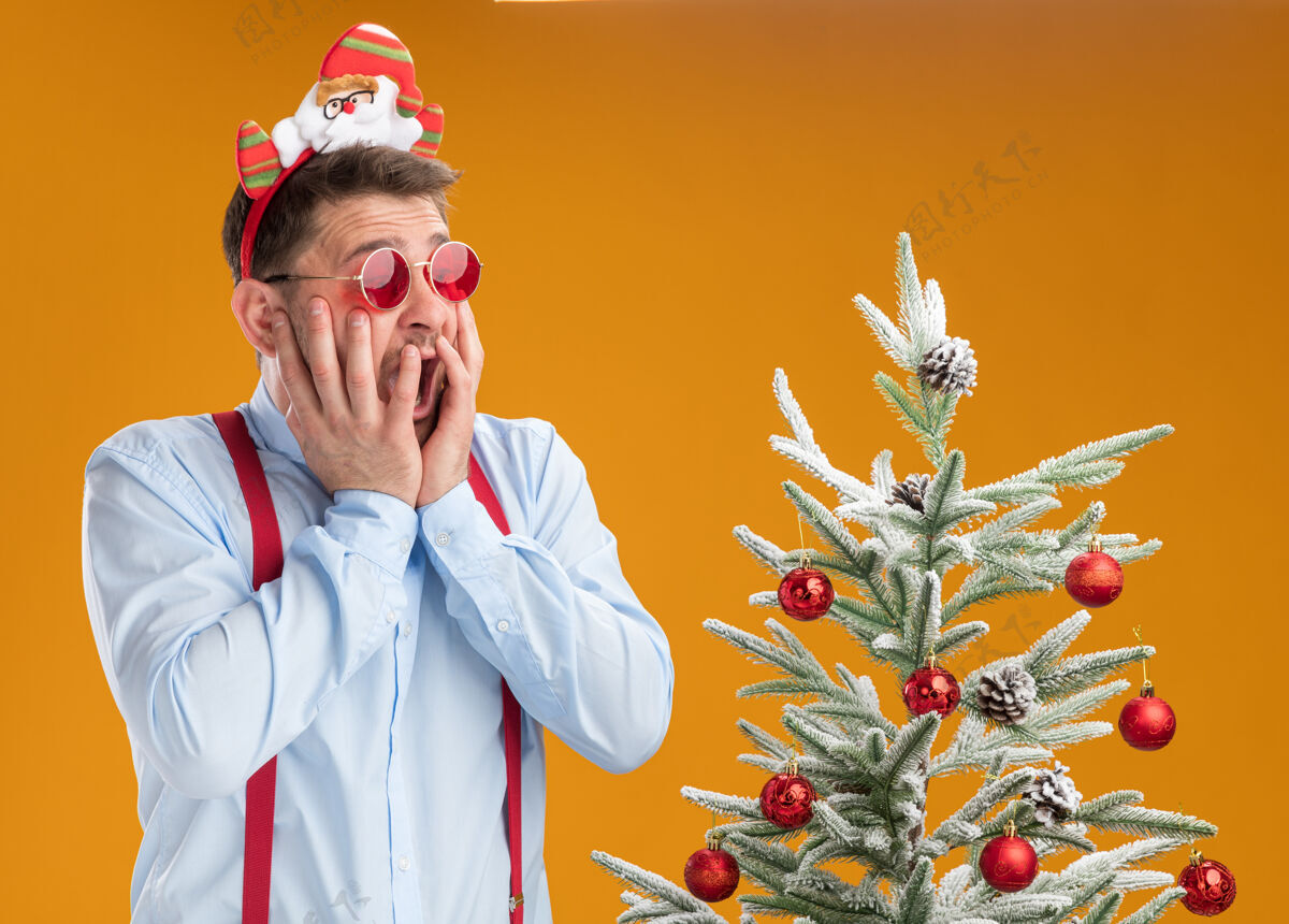 年轻戴着吊带领结的年轻人站在圣诞树旁 戴着圣诞老人和红眼镜 惊恐地看着橙色背景下的圣诞树边缘吊带圣诞树