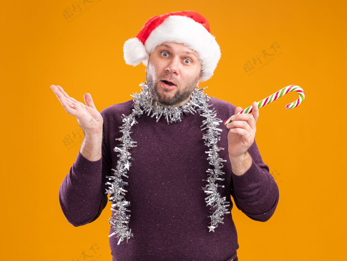 手笨手笨脚的中年男子戴着圣诞帽 脖子上戴着金箔花环 手里拿着圣诞甜手杖 看着摄像机 镜头里只剩下一只孤立在橙色背景上的空手不知所措手杖围着