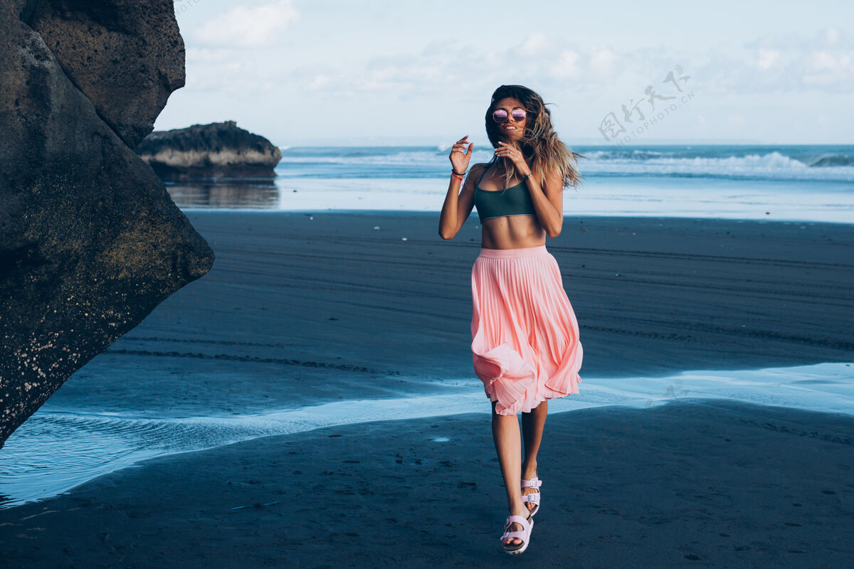 沙滩在黑沙滩上穿着小上衣和粉色裙子的白种人身材的晒黑女人黑沙滩热带健康