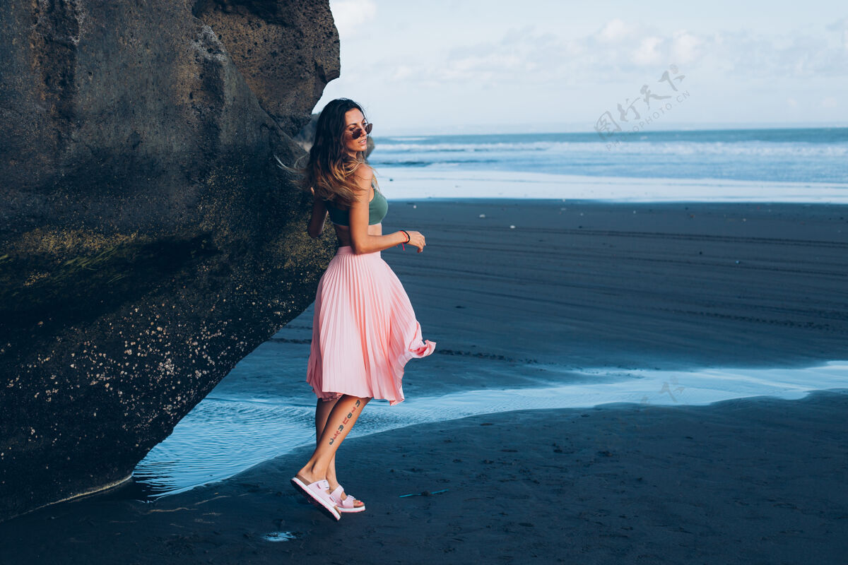 海洋在黑沙滩上穿着小上衣和粉色裙子的白种人身材的晒黑女人户外沙滩女士