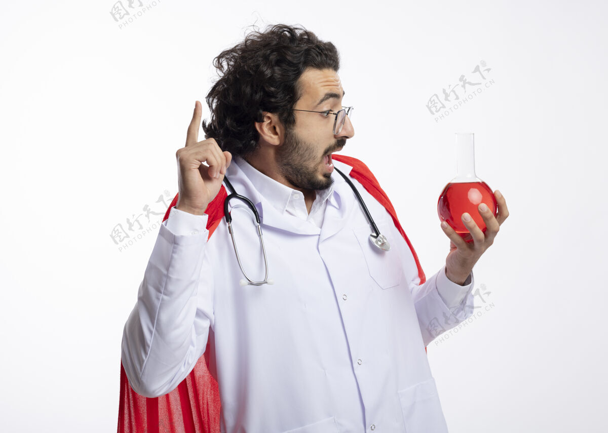 制服一个戴着眼镜 身穿医生制服 披着红色斗篷 脖子上戴着听诊器的年轻白种人惊讶地握着并看着玻璃瓶中的红色化学液体朝上男人斗篷年轻