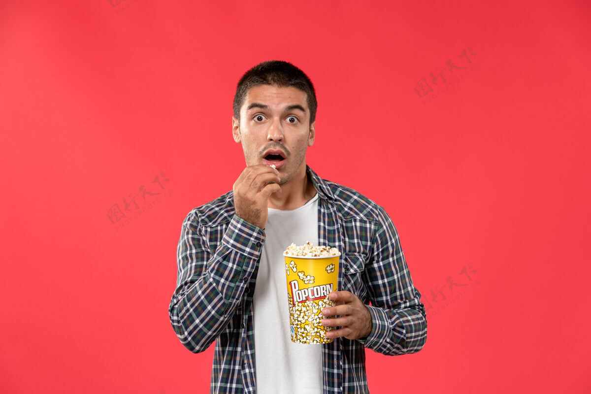 爆米花正面图年轻男子拿着爆米花包在浅红的墙上吃着电影院里的电影男肖像电影电影