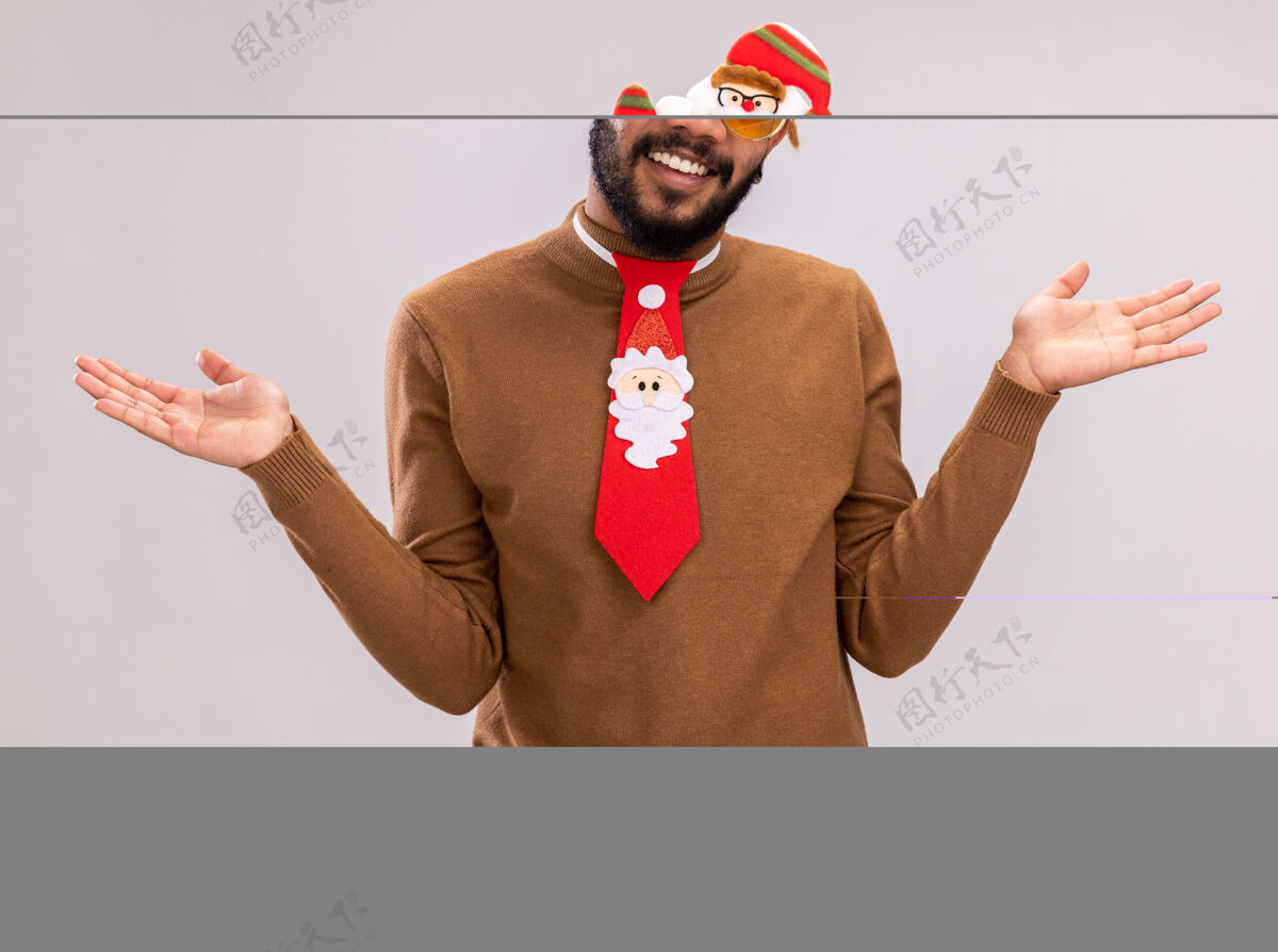 手杖快乐的非洲裔美国人身穿棕色毛衣 头戴圣诞老人戒指 打着有趣的红色领带 手持糖果手杖 微笑着看着摄像机 双臂伸向两侧 站在白色背景上站着头微笑