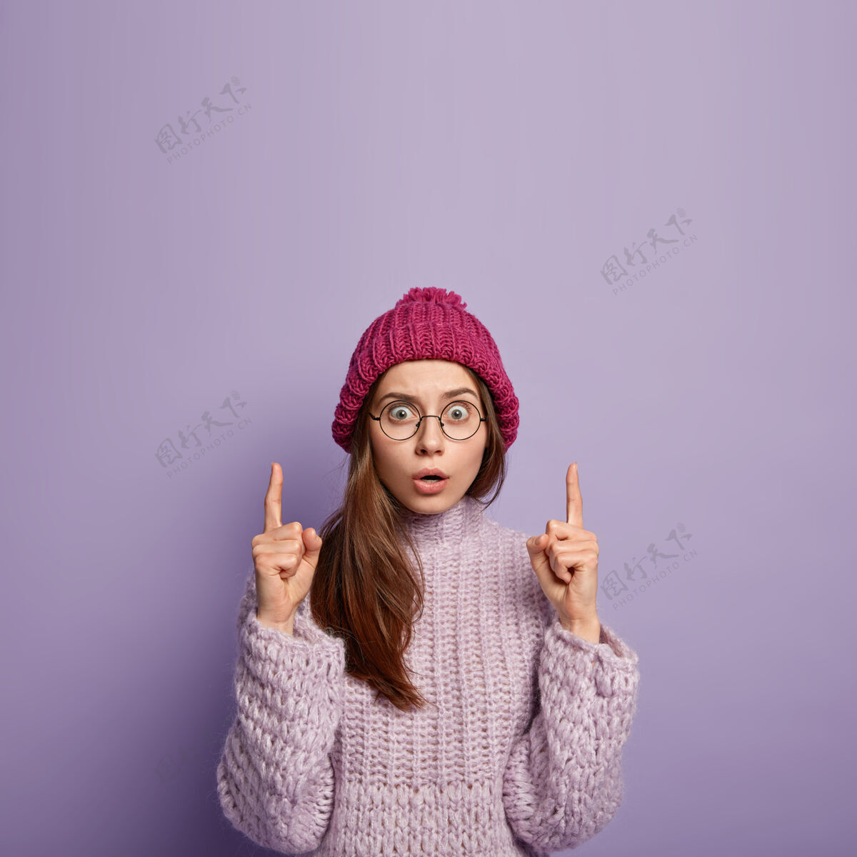 天哪室内拍摄的惊魂未定的年轻白种女性用食指指向上方 戴着透明眼镜 冬衣 隔着紫色的墙壁 自由空间向上帽子演示表情