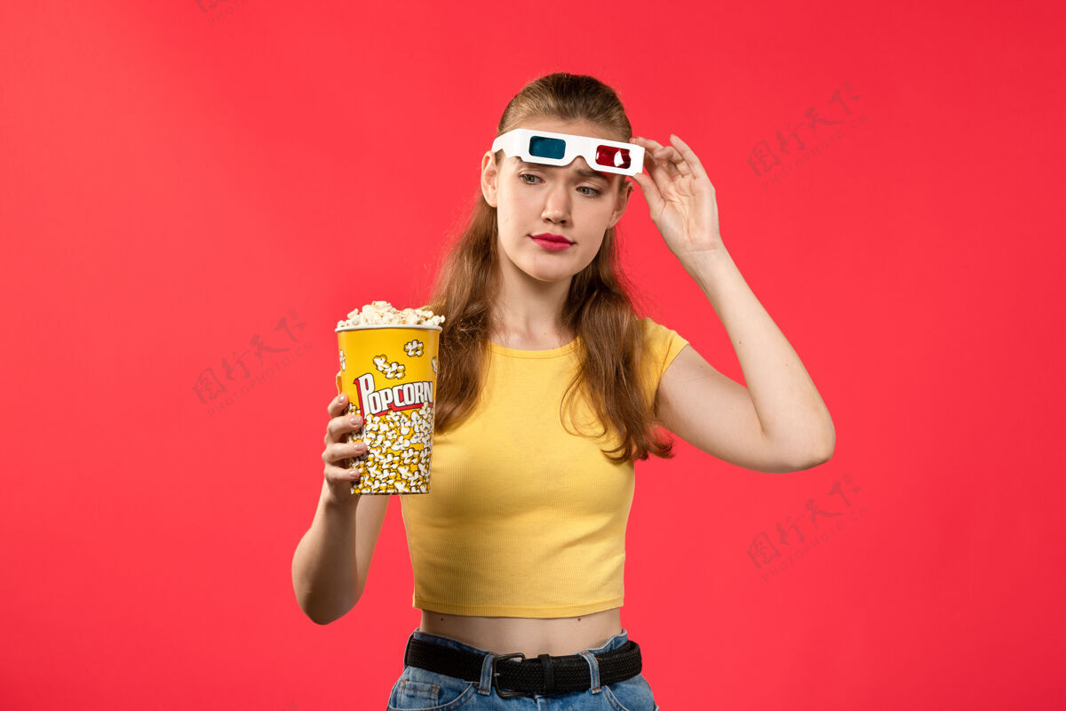 摆姿势正面图年轻女子在电影院拿着爆米花上红墙电影院女彩漂亮女电影