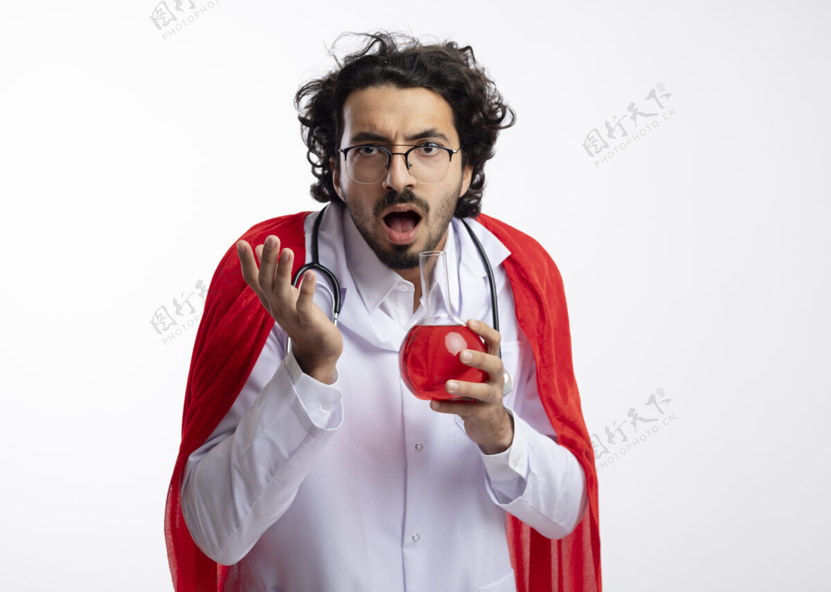 医生无知的白人年轻人戴着眼镜 身穿医生制服 披着红色斗篷 脖子上戴着听诊器 站在那里 举起手来 手里拿着玻璃瓶中的红色化学液体液体持有男人