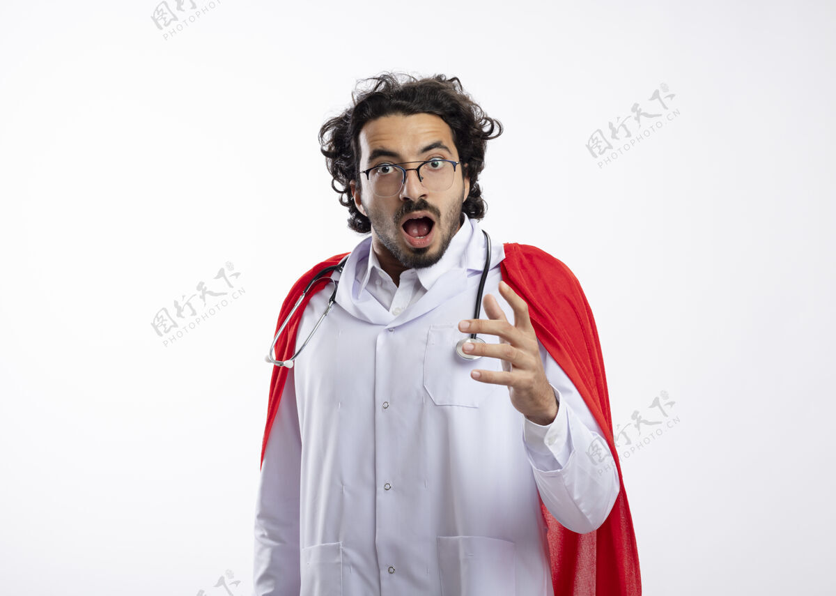 制服震惊的年轻白种人超级英雄戴着眼镜 身穿医生制服 披着红色斗篷 脖子上戴着听诊器 表情和手指眼镜光学周围