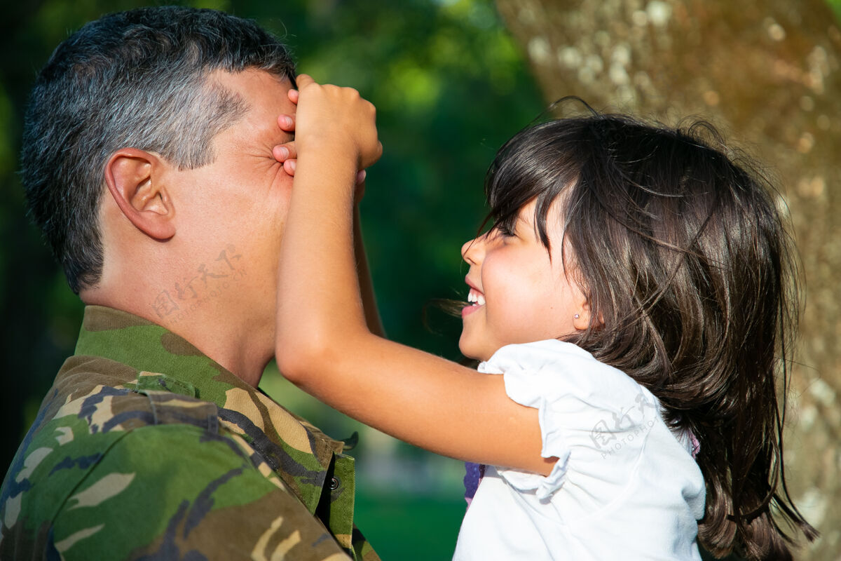 侧视图快乐的军人父亲抱着小女儿 而快乐的女孩则闭上嘴微笑侧视家庭团聚或回家的概念草女儿关闭