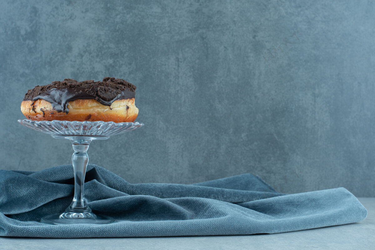 美味甜甜圈放在玻璃基座上 放在大理石上起皱的桌布上巧克力美味烘焙