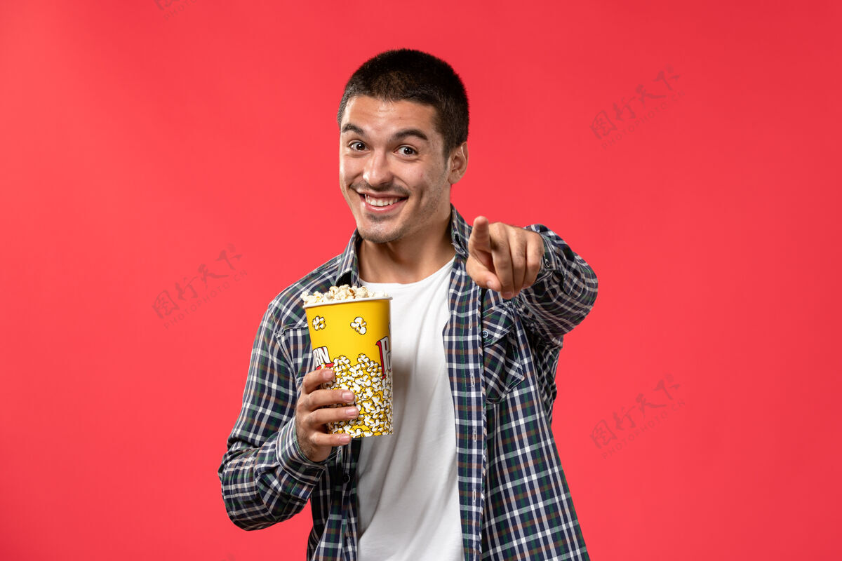 成人正面图年轻男子拿着爆米花包 微笑着站在浅红的墙上看电影的男男孩光年轻电影