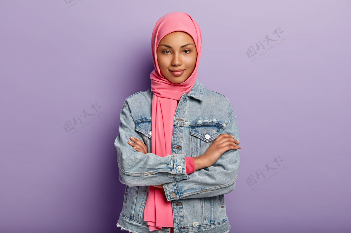 年轻人自信的黑皮肤女性有宗教观 双臂交叉 头戴粉色头巾和牛仔外套 隔着紫色的墙壁 饶有兴趣地听新闻面纱站立高兴
