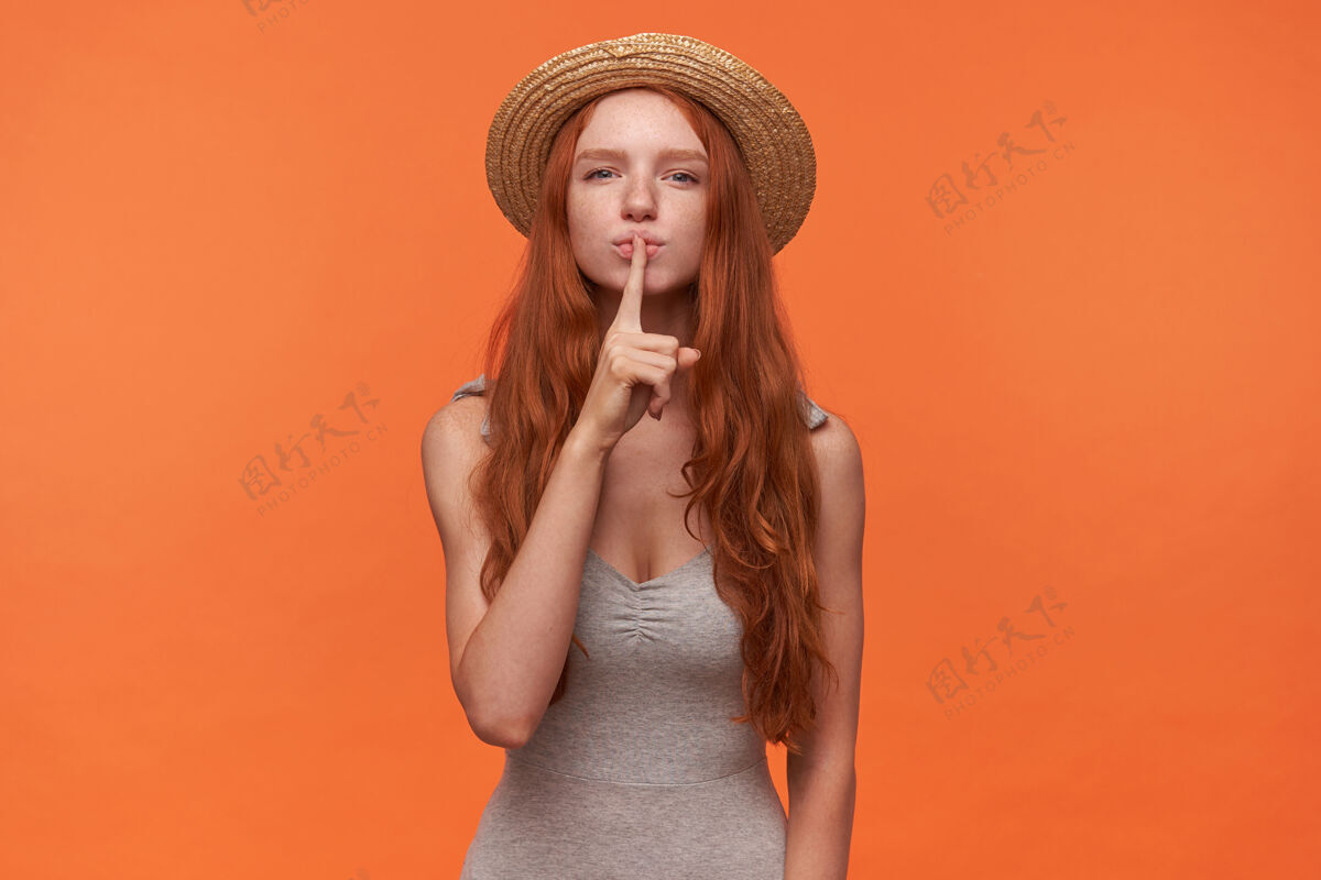 发型一幅迷人的年轻女子的肖像 她卷曲的狐狸精的头发把食指举到嘴唇上 要求保持沉默 穿着灰色衬衫和草帽在橙色背景上摆姿势肖像嘴唇20多岁