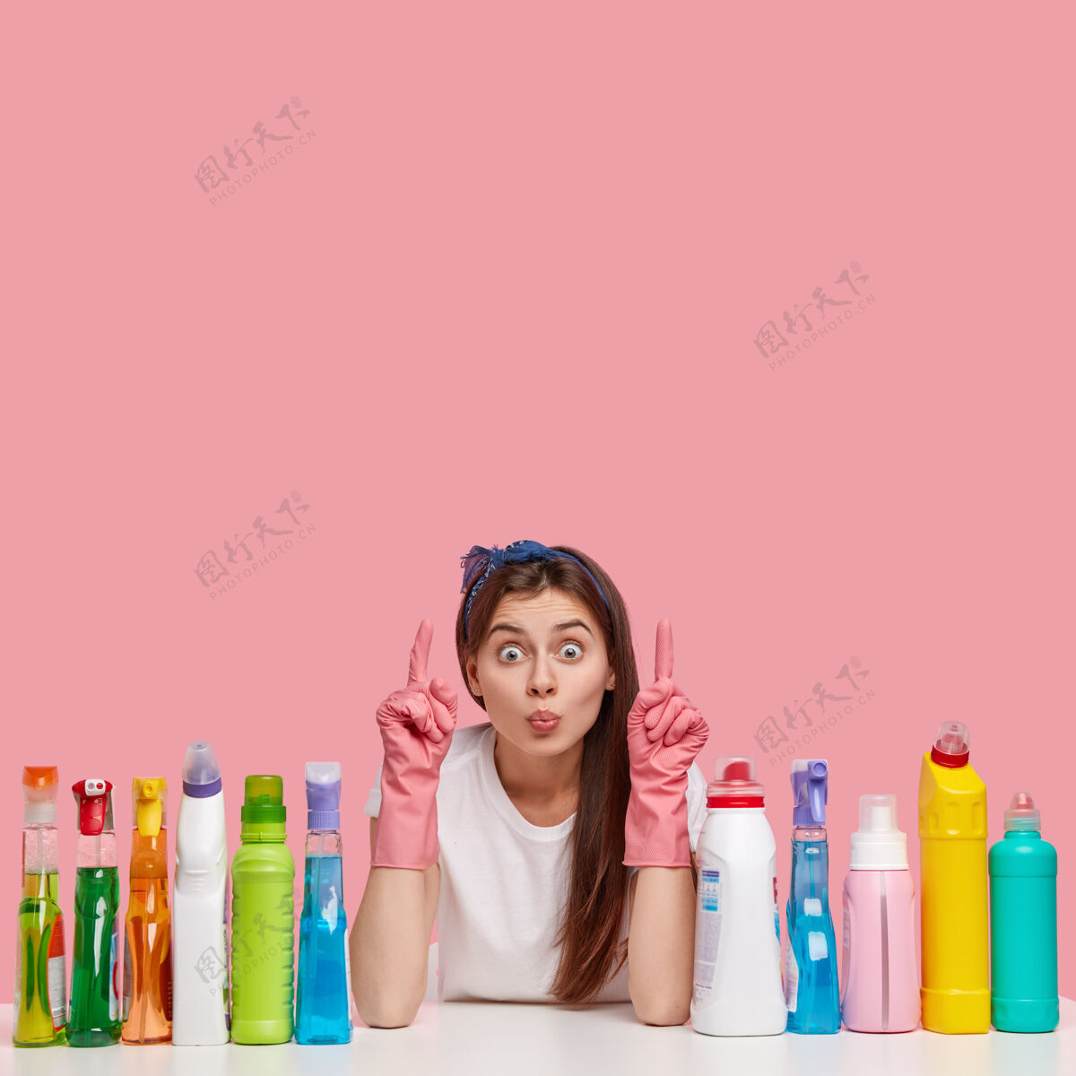 指点迷人女人的垂直镜头保持双唇合拢 指向上方空白处 显示要清洗的地方 周围是各种洗涤剂的瓶子 戴着橡胶防护手套 头带女人清洗清洁剂