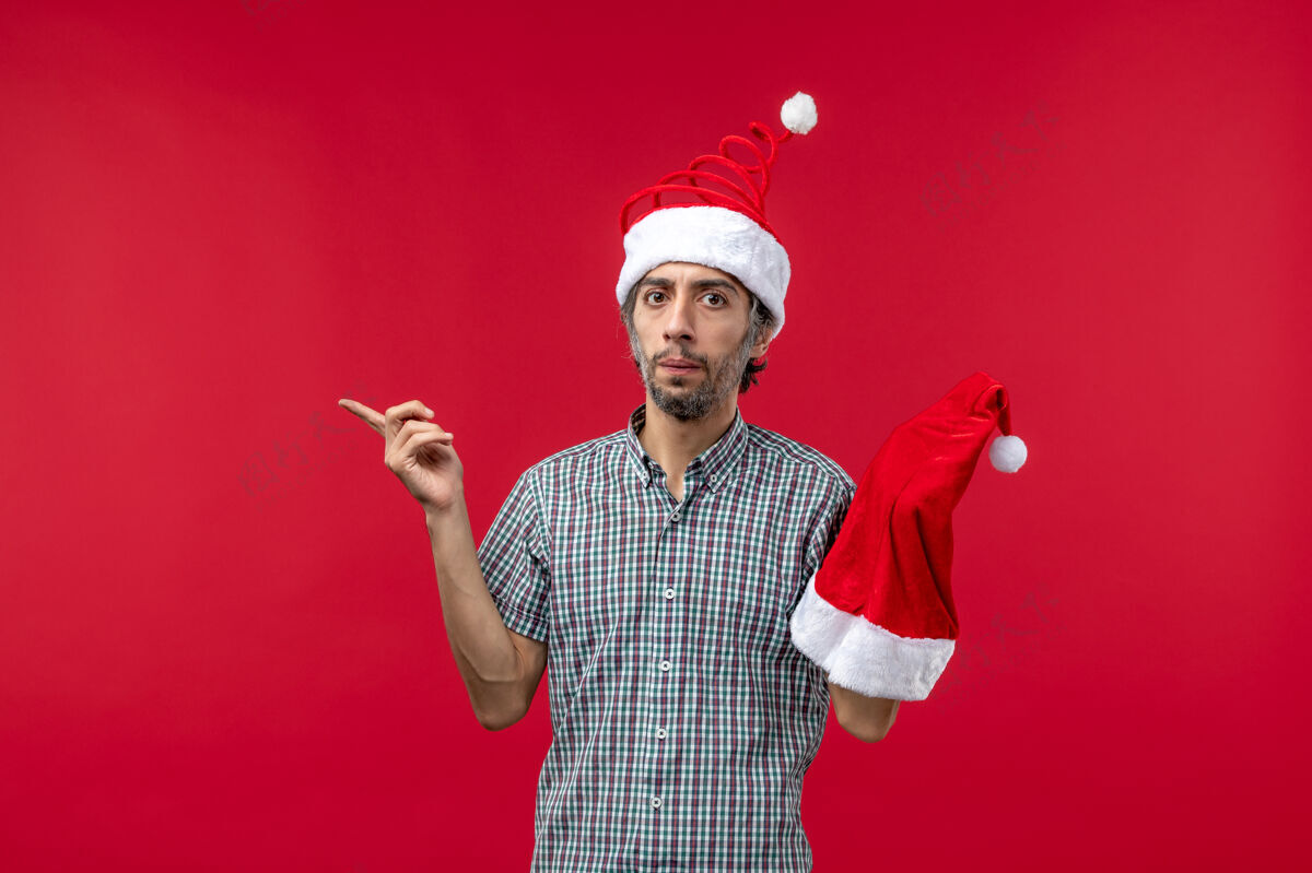圣诞节红墙上拿着圣诞帽的年轻人的正视图帽子成人快乐