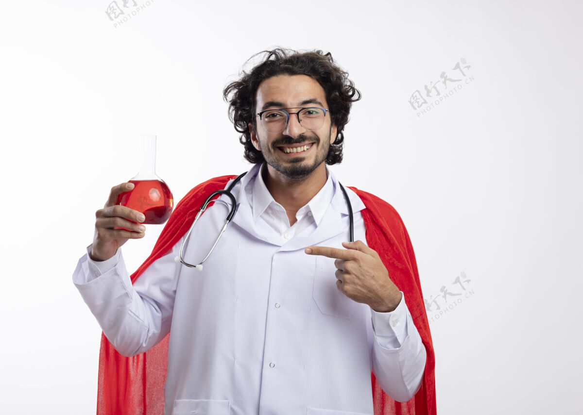 光学一个戴着眼镜 身穿医生制服 头戴红色斗篷 脖子上戴着听诊器 面带微笑的白人年轻人拿着并指着玻璃瓶中的红色化学液体化学微笑液体
