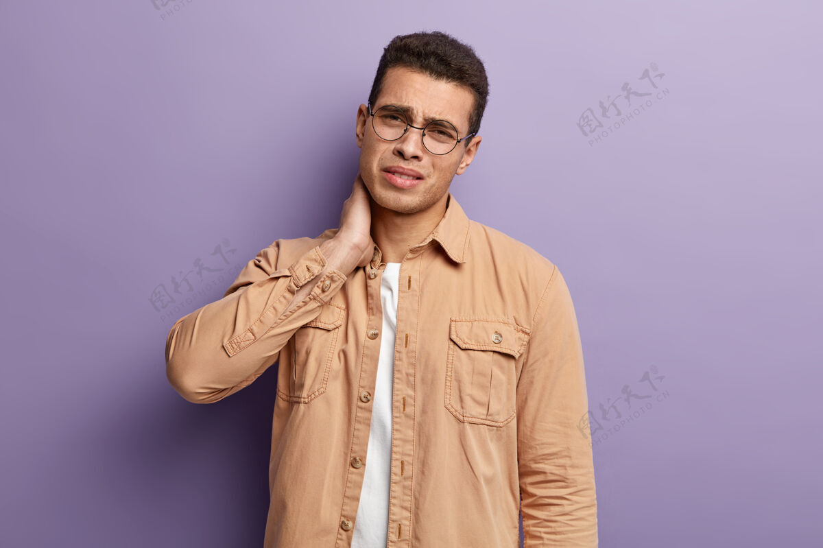 受伤健康问题概念沮丧的年轻白种人颈部疼痛 表情悲伤 穿棕色衬衫 戴圆眼镜 需要按摩 歪着头 隔着紫色的墙痛苦感觉疼痛