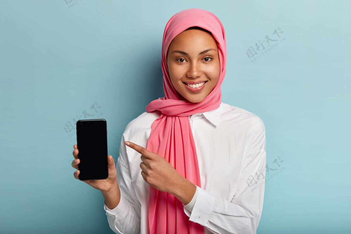 高兴穆斯林女士手持智能手机 显示空白屏幕插入文本或您的信息 戴着粉色头巾和白色衬衫屏幕正面女士